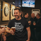 Man in a bar wearing a black t-shirt with humorous text, holding a beer. The man is wearing a Cows Corner t-shirt with a funny darts t-shirt with a slogan 'Throw. Miss. Swear. Repeat'. Cows Corner gifts are perfect for sport-mad fans, these gifts work brilliantly for birthdays, new baby celebrations, Father’s Day, Mother’s Day, Christmas, anniversaries, thank you gifts, end-of-season team awards, graduations, retirements, and just-because moments when you want to raise a smile. 

