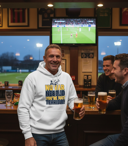 Man in a bar wearing a hoodie with a humorous message, holding a beer, with a TV showing a sports game in the background. The man is wearing a Cows Corner hoodie with the funny football slogan 'No VAR Needed You’re Just Terrible'. Cows Corner gifts are perfect for sport-mad fans, these gifts work brilliantly for birthdays, new baby celebrations, Father’s Day, Mother’s Day, Christmas, anniversaries, thank you gifts, end-of-season team awards, graduations, retirements, and just-because moments 