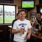 Man in a pub wearing a Cows Corner T-shirt with the funny football slogan on th  front that reads 'Dribble You Can Barely Walk Straight!'   Cows Corner gifts are perfect for sport-mad fans, these gifts work brilliantly for birthdays, new baby celebrations, Father’s Day, Mother’s Day, Christmas, anniversaries, thank you gifts, end-of-season team awards, graduations, retirements, and just-because moments when you want to raise a smile. Designed for people who appreciate thoughtful gifting with a playful edge,