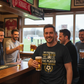 Group of men in a bar watching a football match on television, holding drinks.  The man is wearing a Cows Corner t-shirt with the football slogan on the front that reads 'My Football Coach Is the Best in the World, I’d Know I’ve Played Under the Rest'. Cows Corner gifts are perfect for sport-mad fans, these gifts work brilliantly for birthdays, new baby celebrations, Father’s Day, Mother’s Day, Christmas, anniversaries, thank you gifts, end-of-season team awards, graduations, retirements, and just-because
