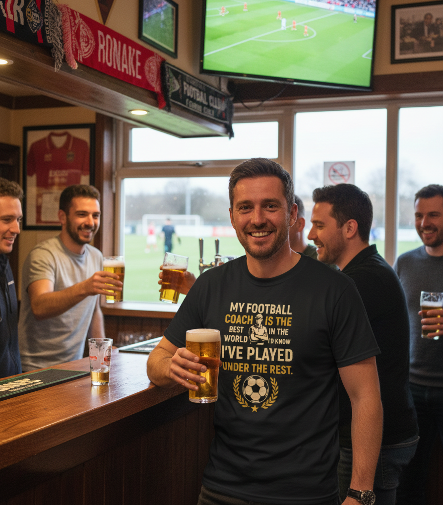 Group of men in a bar watching a football match on television, holding drinks.  The man is wearing a Cows Corner t-shirt with the football slogan on the front that reads 'My Football Coach Is the Best in the World, I’d Know I’ve Played Under the Rest'. Cows Corner gifts are perfect for sport-mad fans, these gifts work brilliantly for birthdays, new baby celebrations, Father’s Day, Mother’s Day, Christmas, anniversaries, thank you gifts, end-of-season team awards, graduations, retirements, and just-because
