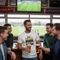 Group of men in a pub watching sports on TV and drinking beer.  The man is wearing a Cows Corner T-shirt with the funny football slogan on the front that reads 'No VAR Needed You’re Just Terrible'.  Cows Corner gifts are perfect for sport-mad fans, these gifts work brilliantly for birthdays, new baby celebrations, Father’s Day, Mother’s Day, Christmas, anniversaries, thank you gifts, end-of-season team awards, graduations, retirements, and just-because moments when you want to raise a smile. 