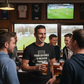 Group of men in a pub watching sports on television and drinking beer.  The man is wearing a Cows Corner T-shirt with the funny football slogan on the front that reads 'Your Defence Is Weaker Than My Grandma’s Wifi'.  Cows Corner gifts are perfect for sport-mad fans, these gifts work brilliantly for birthdays, new baby celebrations, Father’s Day, Mother’s Day, Christmas, anniversaries, thank you gifts, end-of-season team awards, graduations, retirements, and just-because moments when you want to raise