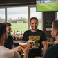 Group of men in a pub watching sports on TV with drinks. The man is wearing a Cows Corner T-shirt with the funny football slogan on the front that reads 'Your First Touch Is a Crime Against Football'. Cows Corner gifts are perfect for sport-mad fans, these gifts work brilliantly for birthdays, new baby celebrations, Father’s Day, Mother’s Day, Christmas, anniversaries, thank you gifts, end-of-season team awards, graduations, retirements, and just-because moments when you want to raise a smile. 