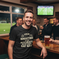 Man in a bar wearing a humorous t-shirt with a sports theme, holding a beer.  The man is wearing a Cows Corner T-shirt with the funny football slogan on the front that reads 'Your First Touch Is a Crime Against Football'. Cows Corner gifts are perfect for sport-mad fans, these gifts work brilliantly for birthdays, new baby celebrations, Father’s Day, Mother’s Day, Christmas, anniversaries, thank you gifts, end-of-season team awards, graduations, retirements, and just-because moments 