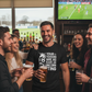 Group of people in a bar watching a sports game on TV, one person wearing a humorous t-shirt.  The man is wearing a Cows Corner T-shirt with the funny football slogan on the front that reads 'Your Tekkers Are As Rare as a Unicorn Sighting'.  Cows Corner gifts are perfect for sport-mad fans, these gifts work brilliantly for birthdays, new baby celebrations, Father’s Day, Mother’s Day, Christmas, anniversaries, thank you gifts, end-of-season team awards, graduations, retirements, and just-because moments