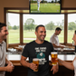 Three people in a bar watching golf on TV, one wearing a humorous t-shirt. The man is wearing a Cows Corner t-shirt with the funny golf slogan One Swing, One Hole, One Very Smug Golfer'. Cows Corner gifts are perfect for sport-mad fans, these gifts work brilliantly for birthdays, new baby celebrations, Father’s Day, Mother’s Day, Christmas, anniversaries, thank you gifts, end-of-season team awards, graduations, retirements, and just-because moments when you want to raise a smile. 
