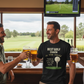Three men in a bar with drinks, smiling and looking at a screen showing a golf course.  The man is wearing a Cows Corner t-shirt with the golf coach slogan on the front that reads 'Best Golf Coach in the World. Thanks for Every Swing, Smile and Slice Fix'. Cows Corner gifts are perfect for sport-mad fans, these gifts work brilliantly for birthdays, new baby celebrations, Father’s Day, Mother’s Day, Christmas, anniversaries, thank you gifts, end-of-season team awards, graduations, retirements
