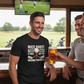 Man wearing a black t-shirt with a humorous golf-themed design, holding a beer, in a bar setting with a TV showing a golf game. The man is wearing a Cows Corner t-shirt with the funny golf slogan on the front that reads 'Nice Shot! Too Bad It Was on the Wrong Fairway'. Cows Corner gifts are perfect for sport-mad fans, these gifts work brilliantly for birthdays, new baby celebrations, Father’s Day, Mother’s Day, Christmas, anniversaries, thank you gifts, end-of-season team awards, graduations, retirements