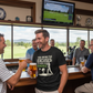 Men enjoying drinks and watching a golf game on TV in a bar. The man is wearing a Cows Corner t-shirt with the funny golf slogan on the front that reads 'You Bring the Excuses, I’ll Bring the Birdies'. Cows Corner gifts are perfect for sport-mad fans, these gifts work brilliantly for birthdays, new baby celebrations, Father’s Day, Mother’s Day, Christmas, anniversaries, thank you gifts, end-of-season team awards, graduations, retirements, and just-because moments when you want to raise a smile. 