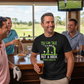 Man in a bar wearing a black t-shirt with a humorous golf-themed design, holding a beer. The man is wearing a Cows Corner t-shirt with a funny golf saying on the front that reads 'You Can Talk to a Fade but a Hook Won’t Listen'. Cows Corner gifts are perfect for sport-mad fans, these gifts work brilliantly for birthdays, new baby celebrations, Father’s Day, Mother’s Day, Christmas, anniversaries, thank you gifts, end-of-season team awards, graduations, retirements, and just-because moments