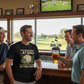 Four men in a bar watching golf on TV, one holding a beer.  The man is wearing a Cows Corner t-shirt with a funny golf slogan on the front that reads 'You’d Be Great at Golf if It Wasn’t for the Hitting Part'. Cows Corner gifts are perfect for sport-mad fans, these gifts work brilliantly for birthdays, new baby celebrations, Father’s Day, Mother’s Day, Christmas, anniversaries, thank you gifts, end-of-season team awards, graduations, retirements, and just-because moments when you want to raise a smile.