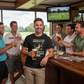 Group of men enjoying drinks at a bar with a golf game on TV in the background. The man is wearing a Cows Corner t-shirt with a funny golf slogan on the front that reads 'Your Golf Game Bring Me Joy, Because It’s Worse Than Mine'.  Cows Corner gifts are perfect for sport-mad fans, these gifts work brilliantly for birthdays, new baby celebrations, Father’s Day, Mother’s Day, Christmas, anniversaries, thank you gifts, end-of-season team awards, graduations, retirements, and just-because moments 