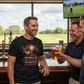 Three men in a bar with one wearing a black t-shirt with a graphic design, holding drinks and smiling.  The man is wearing a Cows Corner t-shirt with the funny golf slogan on the front that reads 'Your Swing Is Like a Horror Movie, Ugly and Full of Screams'. Cows Corner gifts are perfect for sport-mad fans, these gifts work brilliantly for birthdays, new baby celebrations, Father’s Day, Mother’s Day, Christmas, anniversaries, thank you gifts, end-of-season team awards, graduations, retirements