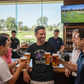 Group of people enjoying beers together in a bar with a golf course view.  The man is wearing a Cows Corner t-shirt with a funny golf slogan on the front that reads 'Your Swing Looks Like a Drunk Octopus Falling out of a Tree'. Cows Corner gifts are perfect for sport-mad fans, these gifts work brilliantly for birthdays, new baby celebrations, Father’s Day, Mother’s Day, Christmas, anniversaries, thank you gifts, end-of-season team awards, graduations, retirements, and just-because moments 