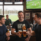 Three men in a pub clinking beer glasses with a television showing a sports match in the background. The man is wearing a Cows Corner t-shirt with the funny rugby slogan 'I’ve Seen Better Hands on a Clock – Phil Kearns'. Cows Corner gifts are perfect for sport-mad fans, these gifts work brilliantly for birthdays, new baby celebrations, Father’s Day, Mother’s Day, Christmas, anniversaries, thank you gifts, end-of-season team awards, graduations, retirements, and just-because moments 