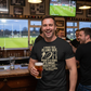 Man in a bar wearing a rugby-themed t-shirt holding a beer, with rugby matches on TV in the background. The man is wearing a Cows Corner t-shirt that has a funny rugby slogan 'A Rugby Tour Is Like Sex. When It’s Good, It’s Great. When It’s Bad, It’s Still Pretty Good – Willie John McBride'. Cows Corner gifts are perfect for sport-mad fans, these gifts work brilliantly for birthdays, new baby celebrations, Father’s Day, Mother’s Day, Christmas, anniversaries, thank you gifts, end-of-season team awards