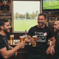 Three men in a pub watching rugby on television, holding beers. The man is wearing a Cows Corner t-shirt with the funny rugby slogan on the front that reads 'If Rugby Was Easy They’d Call It Football'. Cows Corner gifts are perfect for sport-mad fans, these gifts work brilliantly for birthdays, new baby celebrations, Father’s Day, Mother’s Day, Christmas, anniversaries, thank you gifts, end-of-season team awards, graduations, retirements, and just-because moments when you want to raise a smile. 

