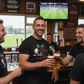 Men enjoying drinks in a pub with a sports game on the TV. The man is wearing a Cows Corner t-shirt with a funny rugby slogan on the front that reads 'No Guts No Glory No Teeth No Problem'. Cows Corner gifts are perfect for sport-mad fans, these gifts work brilliantly for birthdays, new baby celebrations, Father’s Day, Mother’s Day, Christmas, anniversaries, thank you gifts, end-of-season team awards, graduations, retirements, and just-because moments when you want to raise a smile. 

