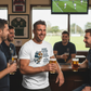 Man in a pub wearing a rugby-themed t-shirt holding a beer, with others around him also drinking. The man is wearing a Cows Corner t-shirt with the funny rugby slogan 'Rugby Players Are Either Piano Movers or Piano Players – Pierre Danos'. Cows Corner gifts are perfect for sport-mad fans, these gifts work brilliantly for birthdays, new baby celebrations, Father’s Day, Mother’s Day, Christmas, anniversaries, thank you gifts, end-of-season team awards, graduations, retirements, and just-because moments 
