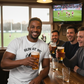 Group of men enjoying drinks and watching a sports match in a pub. The man is wearing a Cows Corner t-shirt with the funny rugby slogan on the front that reads 'Run at Me I Dare You'. Cows Corner gifts are perfect for sport-mad fans, these gifts work brilliantly for birthdays, new baby celebrations, Father’s Day, Mother’s Day, Christmas, anniversaries, thank you gifts, end-of-season team awards, graduations, retirements, and just-because moments when you want to raise a smile. 
