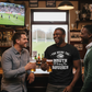 Three men in a pub enjoying drinks and conversation with a sports game on the TV. The man is wearing a Cows Corner t-shirt with a funny Rugby saying on the front that reads 'You Bring the Mouth I’ll Bring the Bruises'. Cows Corner gifts are perfect for sport-mad fans, these gifts work brilliantly for birthdays, new baby celebrations, Father’s Day, Mother’s Day, Christmas, anniversaries, thank you gifts, end-of-season team awards, graduations, retirements, and just-because moments when you want