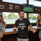 Man in a bar wearing a t-shirt with a humorous message, holding a beer, with people around him.  The man is wearing a Cows Corner T-shirt with the funny tennis slogan on the front that reads 'I Don't Smash Rackets Just Egos'.  Cows Corner gifts are perfect for sport-mad fans, these gifts work brilliantly for birthdays, new baby celebrations, Father’s Day, Mother’s Day, Christmas, anniversaries, thank you gifts, end-of-season team awards, graduations, retirements, and just-because moments.