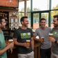 Four men enjoying beers in a sports bar with a tennis-themed shirt and TV screen showing a tennis match.  The man is wearing a Cows Corner T-shirt with the funny tennis slogan on the fron that reads 'Tennis Is My Therapy, Humiliating You Is Just A Bonus'.  Cows Corner gifts are perfect for sport-mad fans, these gifts work brilliantly for birthdays, new baby celebrations, Father’s Day, Mother’s Day, Christmas, anniversaries, thank you gifts, end-of-season team awards, graduations, retirements.