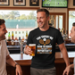 Man in a bar holding a beer, wearing a Cows Corner t-shirt with the tennis coach slogan on the front that reads 'You Taught Me How To Serve but More Importantly How To Believe'. Cows Corner gifts are perfect for sport-mad fans, these gifts work brilliantly for birthdays, new baby celebrations, Father’s Day, Mother’s Day, Christmas, anniversaries, thank you gifts, end-of-season team awards, graduations, retirements, and just-because moments when you want to raise a smile. 