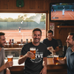 Four men in a bar watching tennis on TV and drinking beer.  The man is wearing a Cows Corner T-shirt with the funny Tennis slogan on the front that reads 'Your Serve Is As Weak As Your Excuses'.  Cows Corner gifts are perfect for sport-mad fans, these gifts work brilliantly for birthdays, new baby celebrations, Father’s Day, Mother’s Day, Christmas, anniversaries, thank you gifts, end-of-season team awards, graduations, retirements, and just-because moments when you want to raise a smile. 