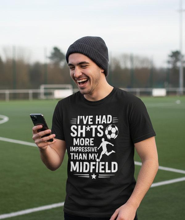 Man on a soccer field wearing a black t-shirt with humorous text and graphics. A Cows Corner t-shirt with the funny football slogan 'I’ve Had Sh*ts More Impressive Than My Midfield'. Cows Corner gifts are perfect for sport-mad fans, these gifts work brilliantly for birthdays, new baby celebrations, Father’s Day, Mother’s Day, Christmas, anniversaries, thank you gifts, end-of-season team awards, graduations, retirements, and just-because moments when you want to raise a smile. 
