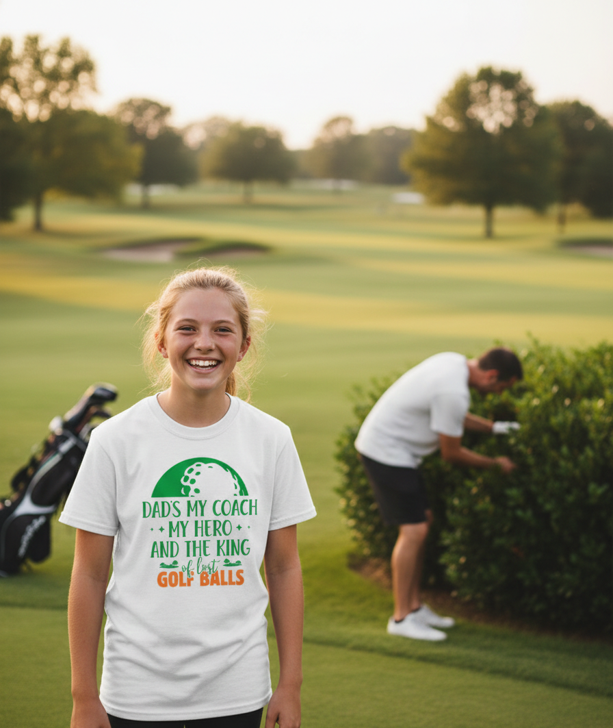 Person wearing a white t-shirt with a golf-themed design on a golf course. The girl is wearing a Cows Corner t-shirt with the funny golf slogan 'Dads My Coach, My Hero and the King of Lost Golf Balls'. Cows Corner gifts are perfect for sport-mad fans, these gifts work brilliantly for birthdays, new baby celebrations, Father’s Day, Mother’s Day, Christmas, anniversaries, thank you gifts, end-of-season team awards, graduations, retirements, and just-because moments when you want to raise a smile. 
