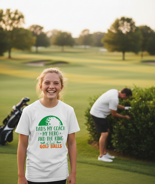 Person wearing a white t-shirt with a golf-themed design on a golf course. The girl is wearing a Cows Corner t-shirt with the funny golf slogan 'Dads My Coach, My Hero and the King of Lost Golf Balls'. Cows Corner gifts are perfect for sport-mad fans, these gifts work brilliantly for birthdays, new baby celebrations, Father’s Day, Mother’s Day, Christmas, anniversaries, thank you gifts, end-of-season team awards, graduations, retirements, and just-because moments when you want to raise a smile. 
