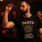 Man holding a dart with a 'Darts Because Therapy is Expensive' t-shirt in a dimly lit room.
