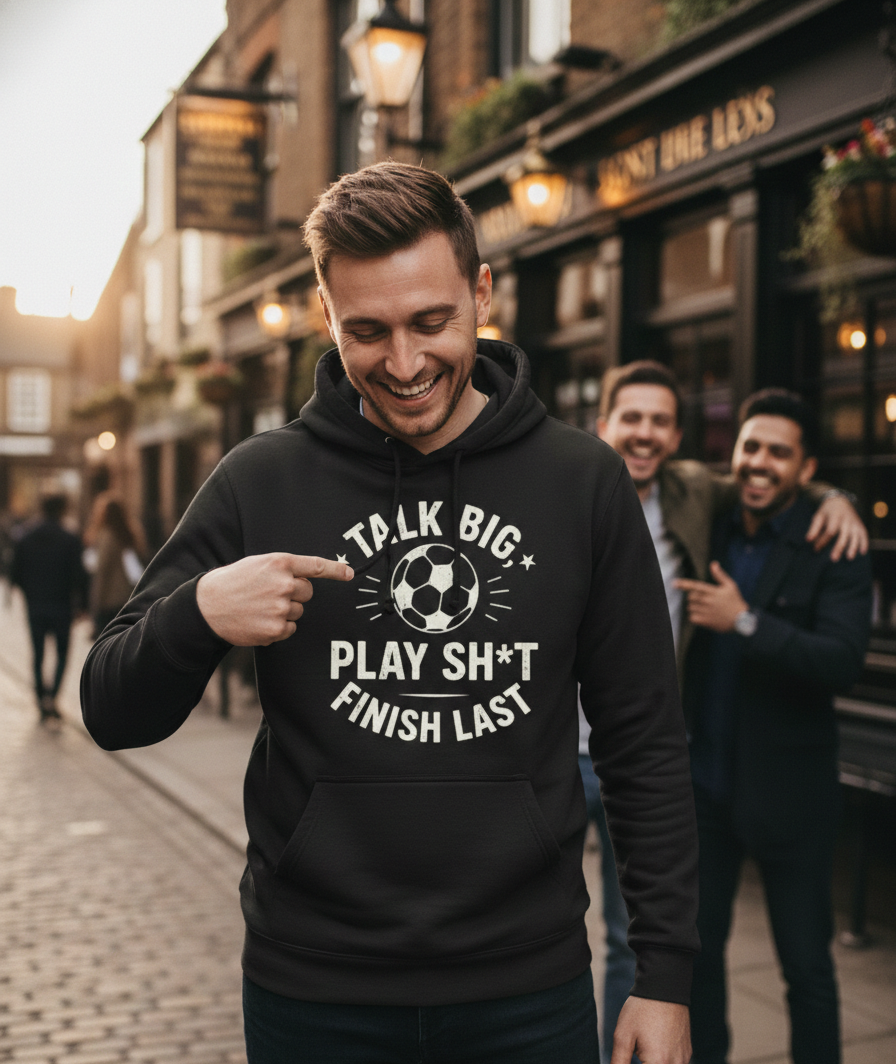 Man wearing a black hoodie with motivational text and pointing at it, standing on a street. The man is wearing a Cows Corner hoodie with the funny football slogan 'Talk Big, Play Sh*t, Finish Last'.  Cows Corner gifts are perfect for sport-mad fans, these gifts work brilliantly for birthdays, new baby celebrations, Father’s Day, Mother’s Day, Christmas, anniversaries, thank you gifts, end-of-season team awards, graduations, retirements, and just-because moments when you want to raise a smile. 
