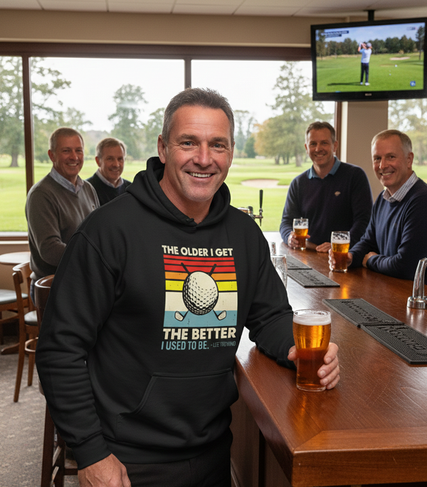 Man wearing a hoodie with a golf-themed design, sitting at a bar with others. The man is wearing a Cows Corner hoodie with a funny golf slogan 'The Older We Get the Better I Used To Be – Lee Trevino'. Cows Corner gifts are perfect for sport-mad fans, these gifts work brilliantly for birthdays, new baby celebrations, Father’s Day, Mother’s Day, Christmas, anniversaries, thank you gifts, end-of-season team awards, graduations, retirements, and just-because moments when you want to raise a smile. 
