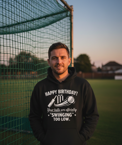 Man wearing a black hoodie with humorous text on a sports field. A Cows Corner hoodie with funny cricket slogan 'Happy Birthday! Your balls are officially 'swinging' too low.' Cows Corner gifts are perfect for sport-mad fans, these gifts work brilliantly for birthdays, new baby celebrations, Father’s Day, Mother’s Day, Christmas, anniversaries, thank you gifts, end-of-season team awards, graduations, retirements, and just-because moments when you want to raise a smile. 
