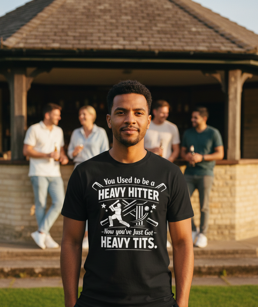 Man wearing a black t-shirt with humorous text in front of a group of people outdoors. A Cows Corner t-shirt with a funny cricket slogan 'You Used to be a Heavy Hitter. Now you’ve Just Got Heavy Tits'.  Cows Corner gifts are perfect for sport-mad fans, these gifts work brilliantly for birthdays, new baby celebrations, Father’s Day, Mother’s Day, Christmas, anniversaries, thank you gifts, end-of-season team awards, graduations, retirements, and just-because moments when you want to raise a smile. 
