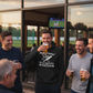 Group of men enjoying drinks and watching TV outside a building. A man wearing a Cows Corner hoodie with the funny cricket birthday slogan on the front that reads 'Happy Birthday! May your wood be firm and your balls stay red. Cows Corner gifts are perfect for sport-mad fans, these gifts work brilliantly for birthdays, new baby celebrations, Father’s Day, Mother’s Day, Christmas, anniversaries, thank you gifts, end-of-season team awards, graduations, retirements, and just-because moments 