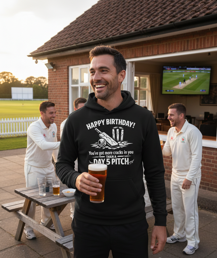 Man wearing a black hoodie with a birthday message, holding a beer, standing outdoors with cricket players and a TV in the background. The man is wearing a Cows Corner hoodie with the funny slogan 'Happy Birthday! You’ve got more cracks in you than a Day 5 pitch'. 