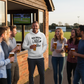 Group of people enjoying drinks outside a pub with a television screen displaying a sports match. The man is wearing a Cows Corner hoodie with the funny cricket birthday slogan 'Old, Bold & Still Looking for a Decent Length. Happy Birthday'. Cows Corner gifts are perfect for sport-mad fans, these gifts work brilliantly for birthdays, new baby celebrations, Father’s Day, Mother’s Day, Christmas, anniversaries, thank you gifts, end-of-season team awards, graduations, retirements, and just-because moments