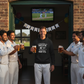 Group of men in cricket uniforms with one holding a beer, standing in front of a television screen showing a cricket match. The man is wearing a Cows Corner hoodie with the funny cricket birthday slogan 'You Used to be a Heavy Hitter. Now you’ve Just Got Heavy Tits.'. Cows Corner gifts are perfect for sport-mad fans, these gifts work brilliantly for birthdays, new baby celebrations, Father’s Day, Mother’s Day, Christmas, anniversaries, thank you gifts, end-of-season team awards, graduations, retirements