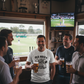 Group of men in a pub watching a sports match on television, holding drinks. The man is wearing a Cows Corner t-shirt with the funny cricket birthday slogan 'Old, Bold & Still Looking for a Decent Length. Happy Birthday'.  Cows Corner gifts are perfect for sport-mad fans, these gifts work brilliantly for birthdays, new baby celebrations, Father’s Day, Mother’s Day, Christmas, anniversaries, thank you gifts, end-of-season team awards, graduations, retirements, and just-because moments 