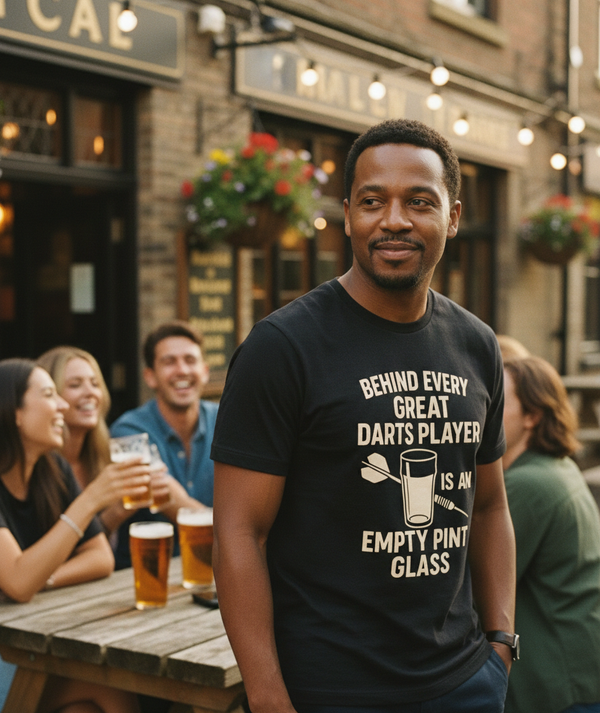 Man wearing a t-shirt with a humorous dart player quote in an outdoor pub setting. A Cows Corner t-shirt with the funny darts slogan 'Behind Every Great Darts Player Is An Empty Pint Glass'. Cows Corner gifts are perfect for sport-mad fans, these gifts work brilliantly for birthdays, new baby celebrations, Father’s Day, Mother’s Day, Christmas, anniversaries, thank you gifts, end-of-season team awards, graduations, retirements, and just-because moments when you want to raise a smile. 
