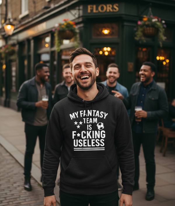 Man wearing a black hoodie with text, standing on a street with friends in the background. The man is wearing a Cows Corner hoodie with the funny football slogan 'My Fantasy Team Is F*cking Useless'. Cows Corner gifts are perfect for sport-mad fans, these gifts work brilliantly for birthdays, new baby celebrations, Father’s Day, Mother’s Day, Christmas, anniversaries, thank you gifts, end-of-season team awards, graduations, retirements, and just-because moments when you want to raise a smile. 
