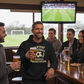 People in a bar watching a soccer game on TV with one person holding a beer.  The man is wearing a Cows Corner hoodie with the funny fantasy football slogan on the front that reads 'Fantasy Football = Weekly Humiliation'. Cows Corner gifts are perfect for sport-mad fans, these gifts work brilliantly for birthdays, new baby celebrations, Father’s Day, Mother’s Day, Christmas, anniversaries, thank you gifts, end-of-season team awards, graduations, retirements, and just-because moments 