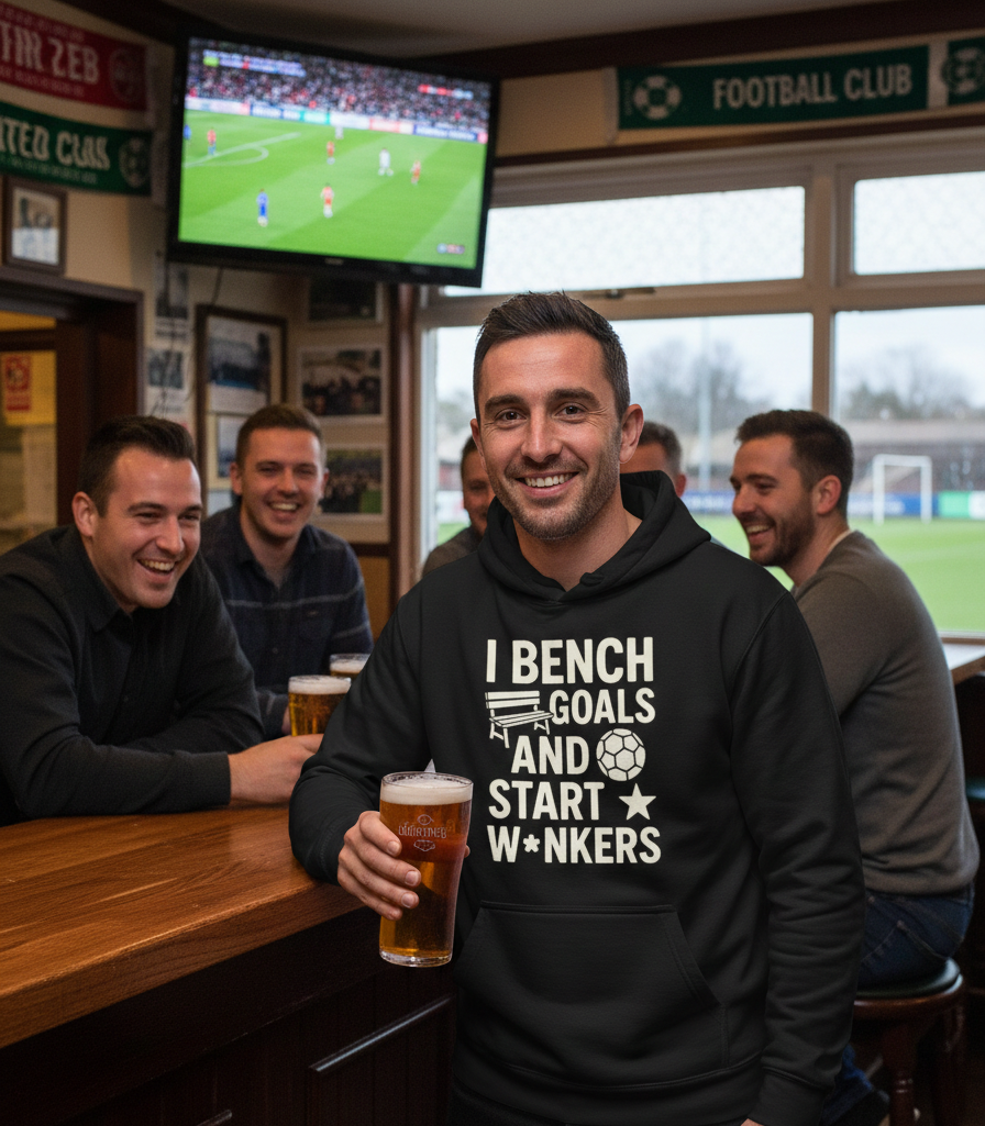 Man in a bar wearing a hoodie with humorous text, holding a beer, with a TV showing a sports game in the background. The man is wearing a Cows Corner hoodie with the funny fantasy football slogan on the front that reads 'I Bench Goals and Start W*nkers'. Cows Corner gifts are perfect for sport-mad fans, these gifts work brilliantly for birthdays, new baby celebrations, Father’s Day, Mother’s Day, Christmas, anniversaries, thank you gifts, end-of-season team awards, graduations, retirements, and just-because