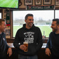 Three men in a pub with a television showing a football match, one wearing a hoodie with humorous text.  The man is wearing a Cows Corner hoodie with the funny fantasy football slogan on the front that reads 'My Fantasy Team Is F*cking Useless'.  Cows Corner gifts are perfect for sport-mad fans, these gifts work brilliantly for birthdays, new baby celebrations, Father’s Day, Mother’s Day, Christmas, anniversaries, thank you gifts, end-of-season team awards, graduations, retirements, and just-because moments