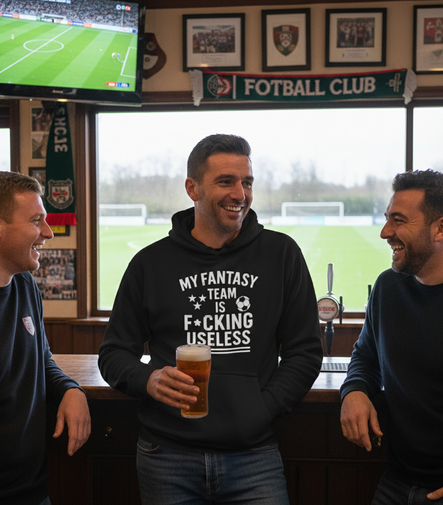 Three men in a pub with a television showing a football match, one wearing a hoodie with humorous text.  The man is wearing a Cows Corner hoodie with the funny fantasy football slogan on the front that reads 'My Fantasy Team Is F*cking Useless'.  Cows Corner gifts are perfect for sport-mad fans, these gifts work brilliantly for birthdays, new baby celebrations, Father’s Day, Mother’s Day, Christmas, anniversaries, thank you gifts, end-of-season team awards, graduations, retirements, and just-because moments