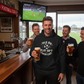 People in a pub watching a football match on TV, with one person holding a beer and wearing a hoodie with text. The man is wearing a Cows Corner hoodie with the funny fantasy football slogan on the front that reads 'Talk Big, Play Sh*t, Finish Last'. Cows Corner gifts are perfect for sport-mad fans, these gifts work brilliantly for birthdays, new baby celebrations, Father’s Day, Mother’s Day, Christmas, anniversaries, thank you gifts, end-of-season team awards, graduations, retirements, and just-because